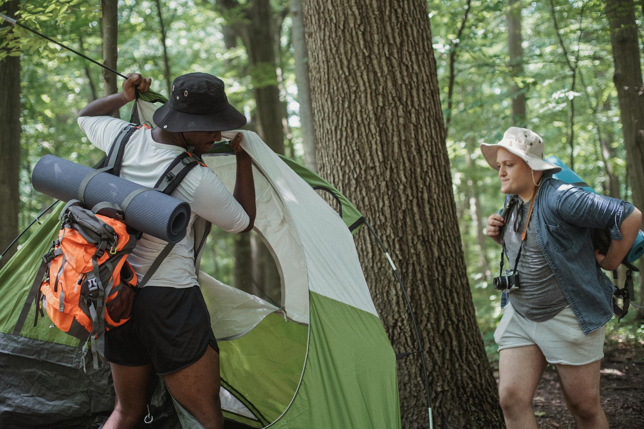 Concentrated young multiracial male hikers in casual clothes and hats with backpacks setting up tent in forest while preparing for recreation during trip