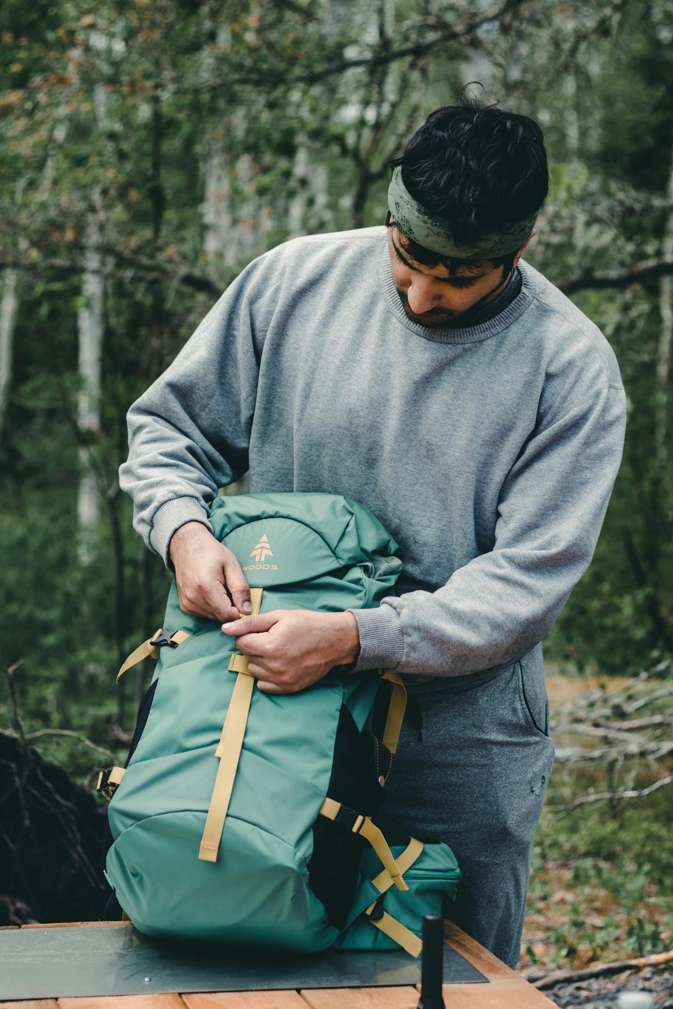Man in gray sweater adjusts green backpack in a lush Canadian forest, showcasing outdoor exploration gear.